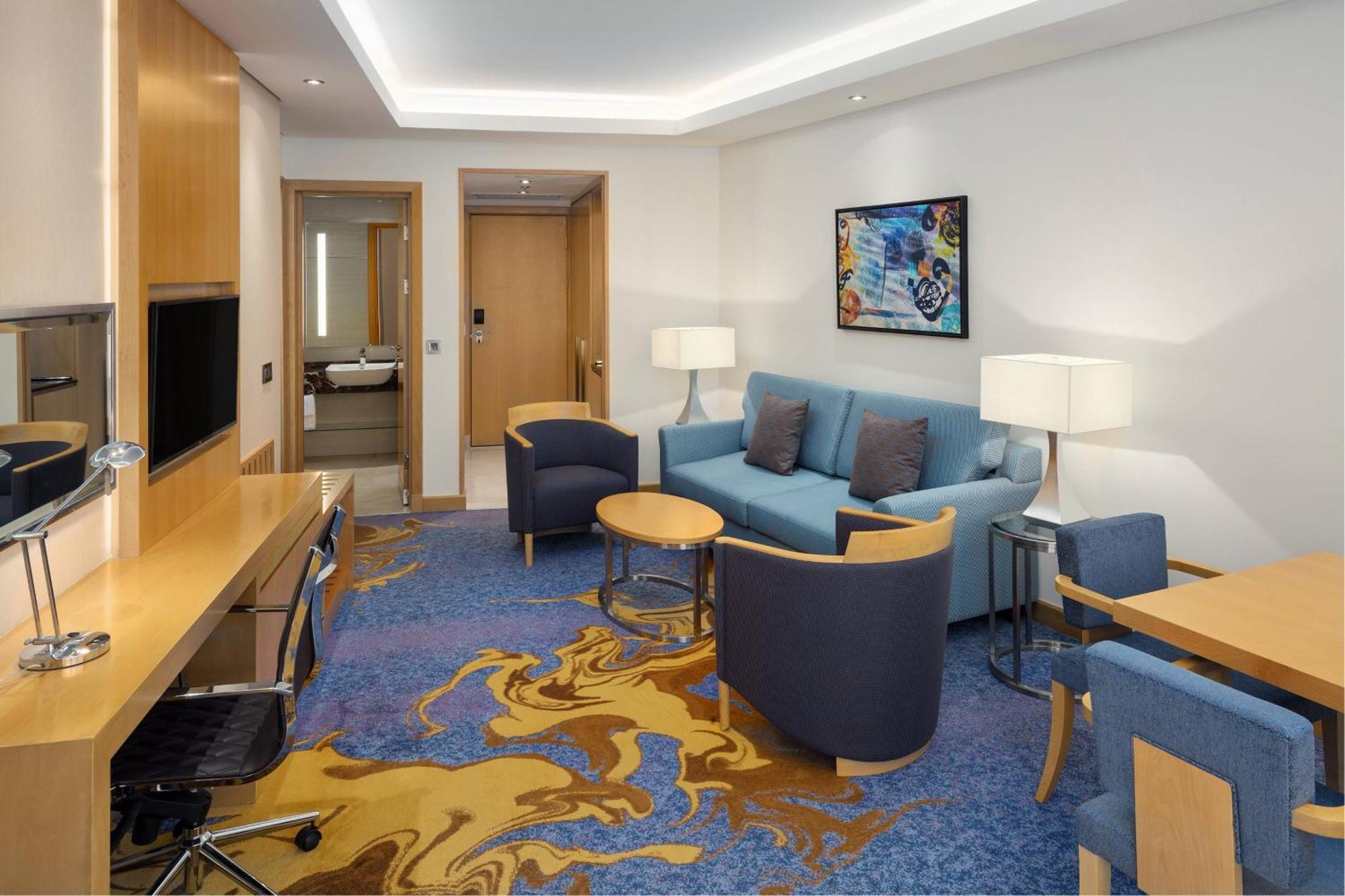 Sheraton Makkah Jabal Al Kaaba Hotel Mecca Екстер'єр фото The photo displays a modern hotel room or suite. The room features a comfortable seating area with a blue couch and two blue armchairs surrounding a small round table. A wooden desk with a black office chair is positioned against one wall, accompanie