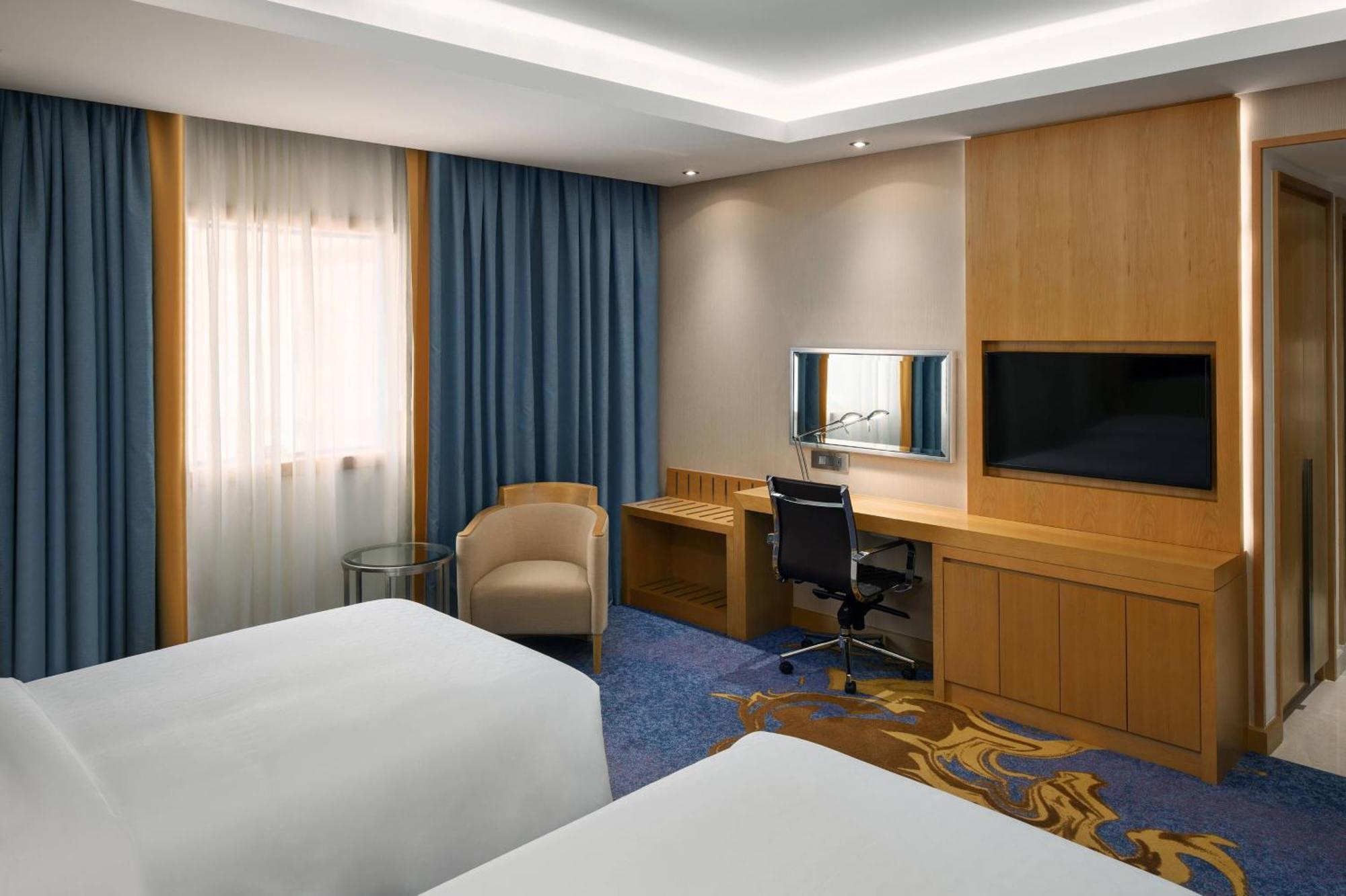Sheraton Makkah Jabal Al Kaaba Hotel Mecca Екстер'єр фото The photo shows a hotel room featuring two beds with white linens. There are large windows covered with blue curtains that allow natural light to enter. On one side of the room, there is a wooden workstation with a chair and a mirror, along with a fl