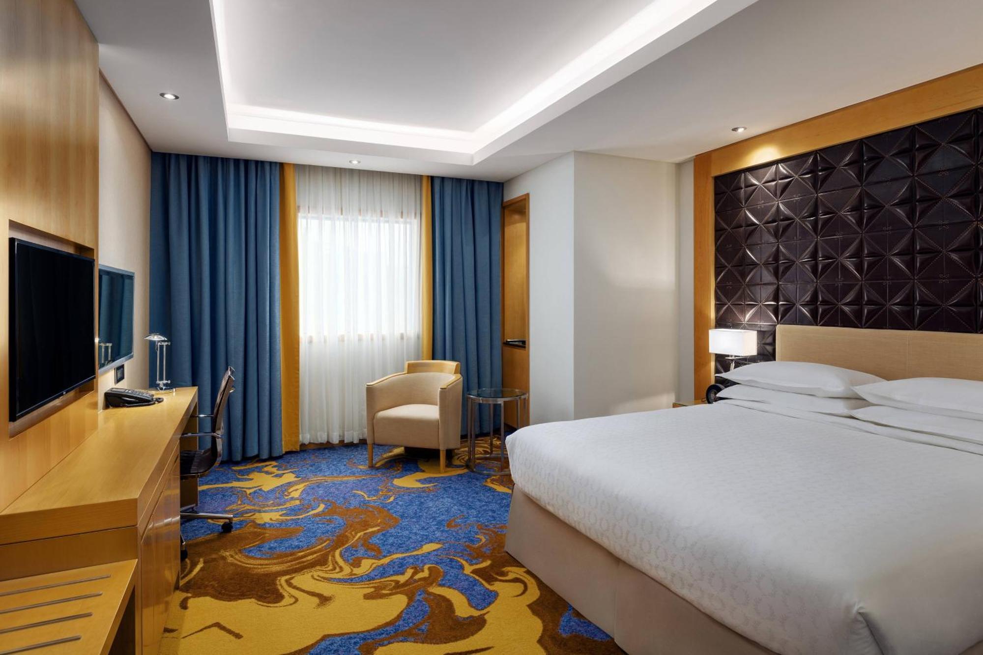 Sheraton Makkah Jabal Al Kaaba Hotel Mecca Екстер'єр фото The photo shows a modern hotel room featuring a comfortable double bed with a light-colored headboard. There's a large window dressed with blue curtains, allowing natural light to enter. The walls are painted in neutral tones, and one wall has a text